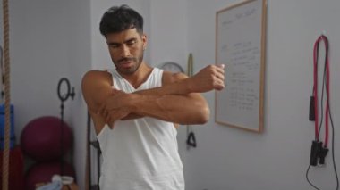 İspanyol bir adam kollarını kapalı bir spor salonunda açıyor, odaklanmış ve kararlı, etrafında spor aletleri ve spor tahtası var..