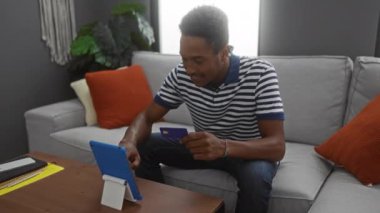 Çizgili gömlekli genç adam, modern oturma odasında tablet ve kredi kartı kullanıyor ve online alışveriş sırasında heyecan gösteriyor.