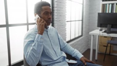 Modern bir ofiste telefonla konuşan genç bir adam, arka planda bir bilgisayarla büyük bir pencerenin yanında oturuyor, profesyonel bir çalışma ortamını aktarıyor..