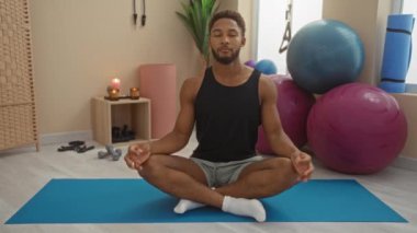 Spor salonundaki mavi yoga minderinin üzerinde meditasyon yapan adam egzersiz ekipmanları ve fitness toplarıyla çevrili.