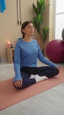 Spor salonunda, yoga minderi, bitki, spor aleti ve arka planda mum ile meditasyon yapan bir kadın..