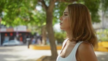 Güneşli bir günde, arka planda yeşil ağaçlar ve şehir manzarasıyla çevrili beyaz tişörtlü bir kadın gülümsüyor ve bir şehir parkında duruyor.