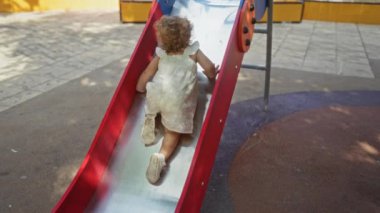 Dışarıdaki bir parkta renkli bir kaydırağın üzerinde oynayan küçük kız güneşli bir günün tadını çıkarıyor, şehir parkında çocukluk masumiyetini ve neşesini gösteriyor.