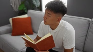 Oturma odasında turuncu yastıklı ve bitkilerle kitap okuyan, ev ortamını tasvir eden ve rahat bir ortamda rahatlamayı veya çalışmayı resmeden genç adam.