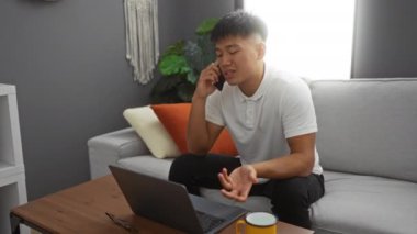 Genç adam modern oturma odasında dizüstü bilgisayar kullanırken telefonla konuşuyor..