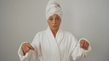 Orta yaşlı bir kadın bornoz ve havlu giyiyor, üzgün ve mutsuz bir ifadeyle işaret ediyor, parmağıyla yön gösteriyor, depresif hissediyor, beyaz arka planda soyutlanıyor.