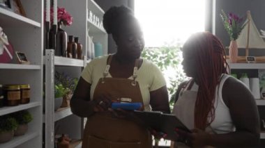 İki Afro-Amerikalı kadın bir ev dekorasyonu mağazasında çeşitli dekorasyon ve bitkilerle çevrili bir tablet ve panoda eşyalar hakkında tartışıyorlar.