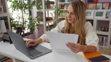 Diz üstü bilgisayarda çalışan sarışın bir kadın arka planda bitki ve raflar olan iç dekorasyon mağazasında kağıtları tutuyor.