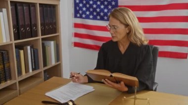 Olgun kadın yargıç, elinde kitap ve belgelerle bir mahkeme salonunda Amerikan bayrağının yanında Birleşik Devletler yargı sistemini temsil ediyor..