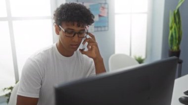 Bilgisayarda çalışan genç bir adam, kapalı alan bitkileri ve mantar mantarlı parlak modern ofis ortamında telefonla konuşuyor..