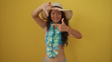 Yaz şapkası takan genç Çinli kadın ve Hawaii lei gülüyor ve elleri ve parmaklarıyla çerçeve yapıyor. Sarı arka planda mutlu bir yaratıcılık ve fotoğraf konsepti fotoğrafı.