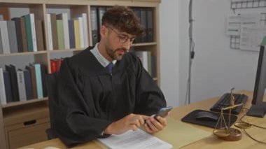 Yargıç cüppeli genç adam, yasal belgeler ve hukuk kitaplarıyla çevrili cep telefonuna bakarken ofiste yumruk tokuşturarak kutlama yapıyor..