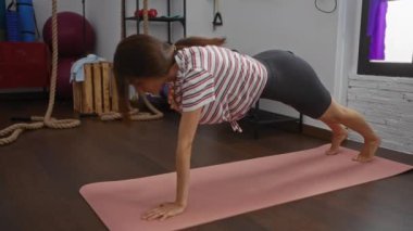 Spor salonunda egzersiz yapan bir kadın. Kapalı alanda paspas üzerinde kalas yapıyor. Rahat çizgili tişört ve şortla spor salonunda egzersiz yapan esmer bir kadın.