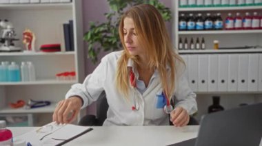 Klinikte oturan kadın doktor, sağlık hizmetleri için profesyonel ve odaklanmış bir atmosfer gösteren tıbbi raflar ve aletlerle çevrili gözlükleri ayarlıyor..