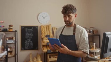 Genç adam bir pastanede çalışırken etrafında taze ekmek ve hamur işleri olan bir tablet kullanıyor.