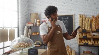 Fırında genç bir adam, etrafında taze ekmek ve hamur işleriyle çevrili dijital tablet kullanırken telefonla konuşuyor..