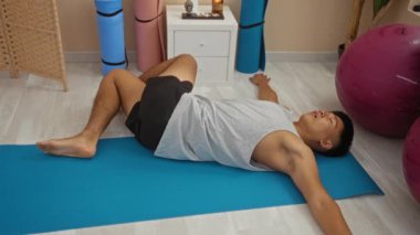 Genç adam, arka planda yoga minderleri ve fitness toplarıyla bir sağlık merkezinde kapalı alanda egzersiz yapıyor.