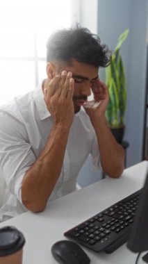 Beyaz gömlekli genç adam, ofiste bitkiler ve bilgisayarla otururken stres ve hayal kırıklığı gösteriyor.