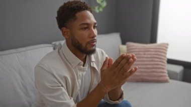 Genç adam modern bir oturma odasında oturuyor, ellerini düşüncelere daldırarak ya da stresle yüzünü kapatıyor.