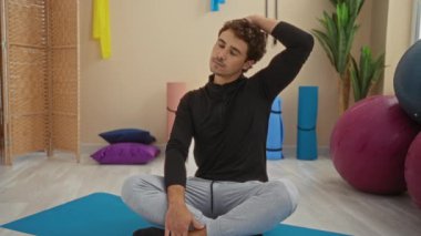 Spor salonunda boyun geren, yoga ekipmanları ve renkli yastıklarla çevrili sakin bir spor merkezinde rahat giyinen bir adam..