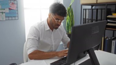 Modern bir ofiste bilgisayar üzerinde çalışan odaklanmış bir adam, verileri analiz ediyor ve arka planda strateji planlaması yaparak notlar yazıyor.