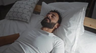 Genç sakallı bir adam beyaz çarşaflar ve yastıklarla evinde loş bir yatak odasında dinleniyor.