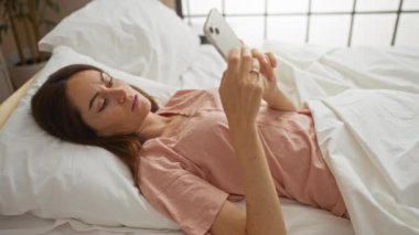 Genç bir kadın, rahat bir yatak odasında yatarken cep telefonuna şaşkın şaşkın bakıyor.