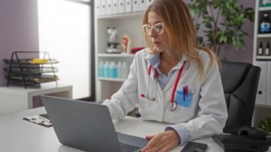 Klinikte dizüstü bilgisayarda çalışan kadın doktor, tıbbi araştırmalara odaklanırken, klinik ekipmanları ile çevrili ve gözlük takıyor..