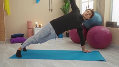 Spor salonunda egzersiz yapan genç bir adam, parlak bir stüdyoda spor topu ve ekipmanla yoga minderinde tahta poz veriyor..