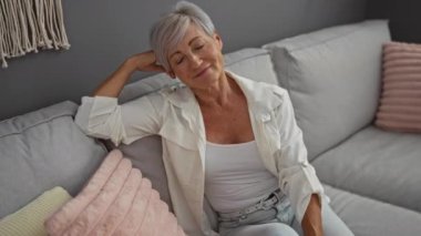 Kısa gri saçlarıyla rahatlayan mutlu bir kadın rahat bir oturma odasında koltukta rahatça oturur.