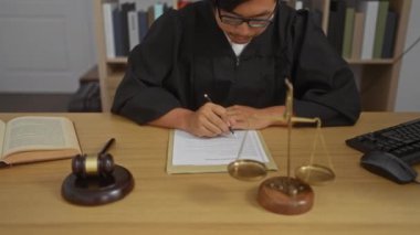 Young man in judge robe signing document at office desk with scales of justice, gavel, and keyboard indicating legal work environment