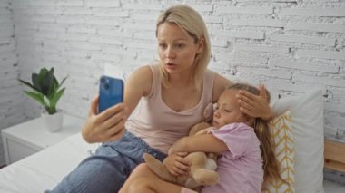 Hasta kızını yatakta rahat ettirirken akıllı telefon kullanıp yatak odasında beyaz tuğladan duvar ve komodinin üzerine bitki yerleştiriyor.