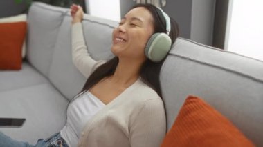 Evdeki kanepede dinlenen genç bir kadın, rahat bir oturma odasında kulaklıkla müziğin tadını çıkarıyor.