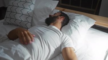 Beyaz bir tişört giymiş, loş bir yatak odasında şablonlu yastıkla uyuyan ve modern bir iç dekorasyonu olan barışçıl bir ifade sergileyen bir adam..
