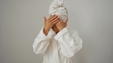 Havlu ve bornoz giymiş orta yaşlı bir kadın, izole edilmiş beyaz arka planda utanmış ve şok olmuş bir ifadeyle parmaklarının arasından dikizliyor.