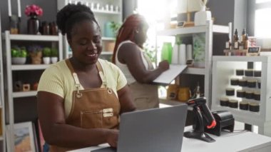 Bir ev dekorasyonu mağazasında çalışan Afrikalı Amerikalı kadınlar, biri dizüstü bilgisayar kullanırken diğeri envanter tutuyor, çeşitli dekoratif eşyalar ve ev eşyaları ile çevrili..