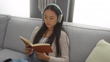 Kadın kitap okuyor ve rahat oturma odasında kulaklıkla müzik dinliyor, rahat bir yastık ve modern dekorla kanepede dinleniyor.