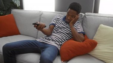 Uzaktan kumandalı bir kanepeye uzanmış parlak yastıkları ve günlük kıyafetleri olan rahat bir oturma odasında rahatlamış bir şekilde duran genç adam..