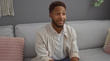 Evdeki bir kanepede oturan genç adam mutlu görünüyor ve modern bir oturma odasında yanlamasına işaret ediyor..