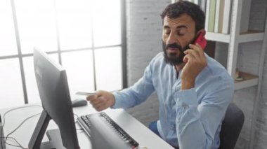 Ofisteki genç adam elinde kredi kartıyla bilgisayar monitörüne bakarken kırmızı bir telefonla konuşuyor.