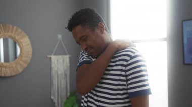 Parlak oturma odası ortamında sırt ağrısı çeken genç bir adam. Çağdaş bir dekor arka planıyla çizgili gömlekle yakalanmış. Evdeki rahatsızlığı gösteriyor..