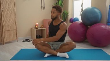 Spor salonunda mavi paspasın üzerinde uzanan genç bir adam. Arka planda egzersiz topları ve mumlarla.