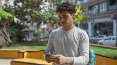 Parlak bir günde akıllı telefon kullanarak şehir merkezinde ağaçlar ve binalarla çevrili bir parkta duran genç adam..