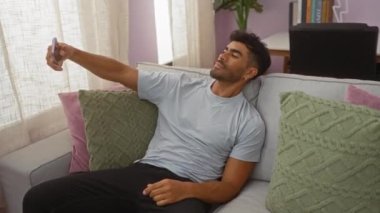 Genç İspanyol bir adam akıllı telefonuyla selfie çekerken rahat bir oturma odasında yastık ve kitaplarla süslenmiş bir kanepede dinleniyor.