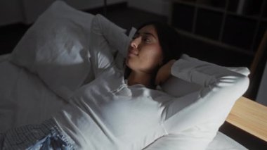 Gece boyunca beyaz bir yatakta yatarken yatak odasında sakin bir ifadeyle dinlenen İspanyol kadın.