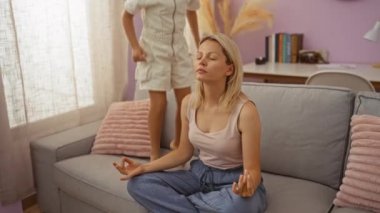 Kızlar parlak oturma odasında pastel dekor ve rahat atmosferle oynarken kadınlar kanepede meditasyon yapıyor.