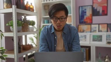 Gözlüklü genç bir adam bir ev dekoratörü dükkanında dizüstü bilgisayara bakıyor saksı bitkileri rafları ve duvarlarda çerçeveli sanat eserleri var.