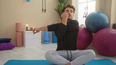 Spor salonundaki bir paspasın üzerinde egzersiz yapan genç adam, etrafını saran spor aletleri ve yoga toplarıyla, huzurlu ve düzenli bir ortamda esneme rutinine odaklanıyor.