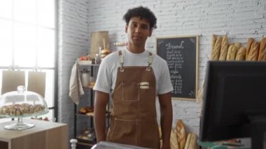 Genç adam bir fırında kolları çapraz duruyor, beyaz duvarlı, iyi aydınlatılmış bir odada ekmek ve hamur işleri sergiliyor, modern bir iç mekanı taze pişmiş malların kırsal cazibesiyle harmanlıyor..