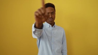 Genç Afro-Amerikalı gömlekli bir adam parmağını kaldırmış jest yapmadan, izole edilmiş sarı arka planda güçlü bir ifade sergiliyor.